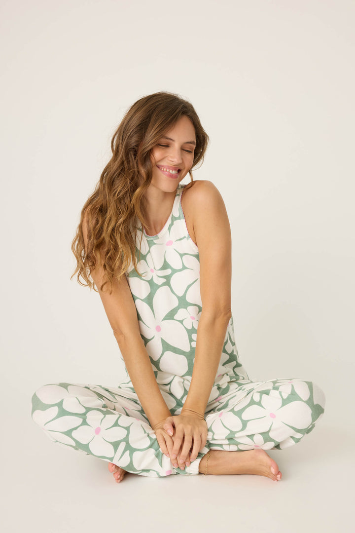 A woman with long wavy hair sits cross-legged, smiling with her eyes closed. She wears P.J. Salvage Wish You Were Here Floral Print Banded Pants in a green and white pattern with pink accents, set against a plain light background.