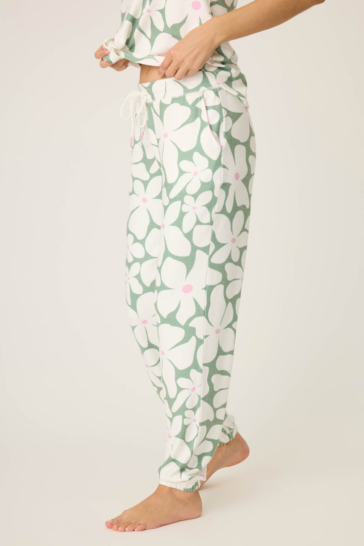 A person stands barefoot, showing only their lower half in P.J. Salvage's Wish You Were Here Floral Print Banded Pants—loose drawstring pants with a green and white floral pattern plus pink accents—against a plain background.