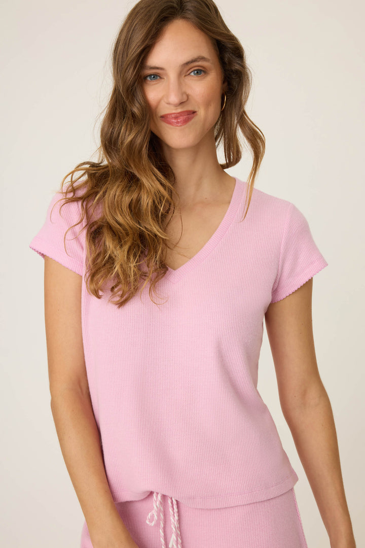 A woman with long wavy brown hair wears the P.J. Salvage Wish You Were Here Peachy Short Sleeve Top in light pink, paired with matching pants. She stands against a plain light background, smiling slightly at the camera.