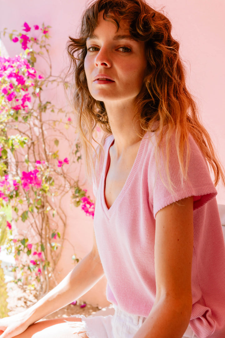 A person with wavy hair wears the P.J. Salvage Wish You Were Here Peachy Short Sleeve Top, sitting indoors near a sunlit window with pink flowers in the background.