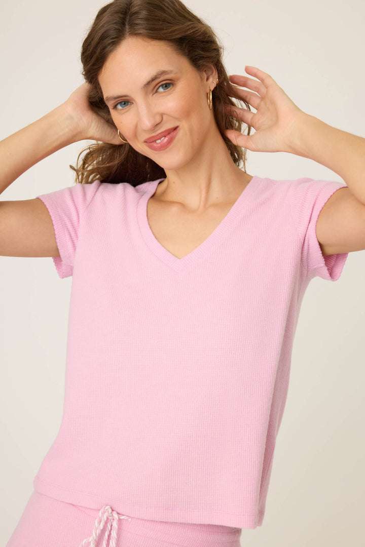 A woman with wavy brown hair wears the P.J. Salvage Wish You Were Here Peachy Short Sleeve Top, standing against a plain background with her hands raised near her head and a slight smile on her face.