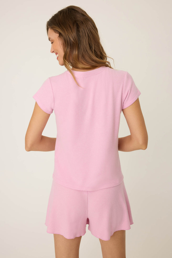 A woman with long brown hair stands with her back to the camera, wearing the P.J. Salvage Wish You Were Here Peachy Short Sleeve Top, in a plain off-white setting.