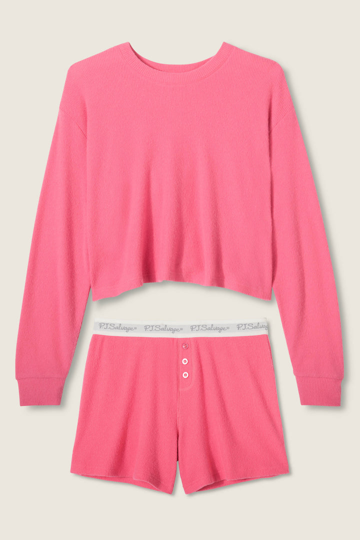The P.J. Salvage Boyfriend Boxer Solid Pink Short Set features a long-sleeve cropped crewneck top and high-waisted shorts with a white elastic logo waistband and three-button front, set against a light beige background.