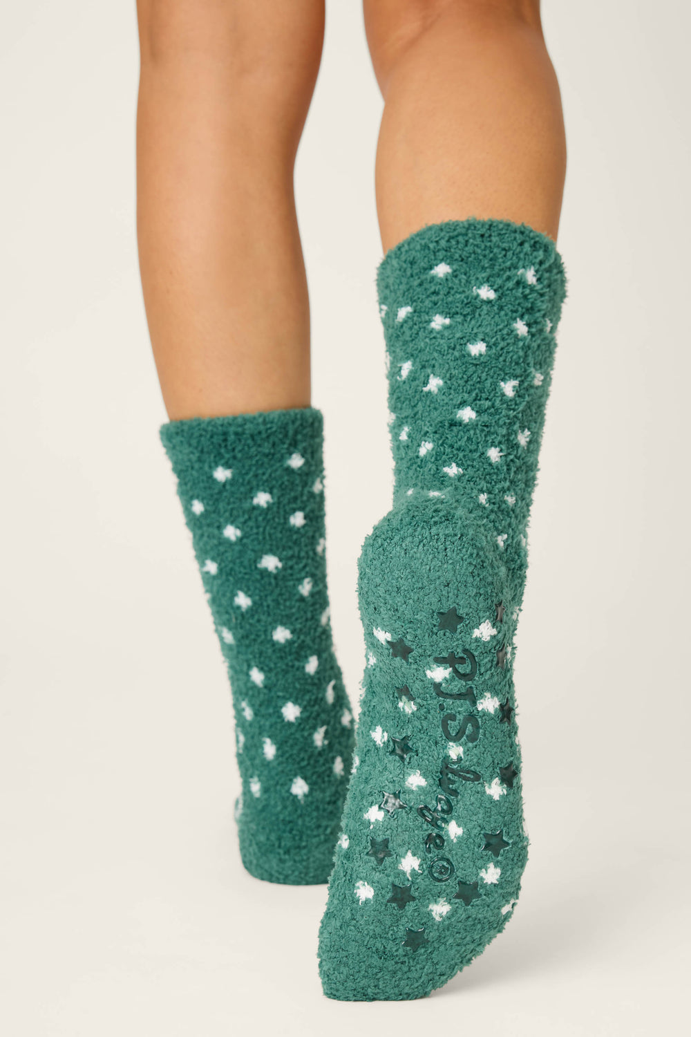 A person wears P.J. Salvage DOTS SOCKS LET'S GET TOASTY—green fuzzy socks with white polka dots—on a light background. The sock’s sole features grips and printed text. Only the lower legs and feet are shown.