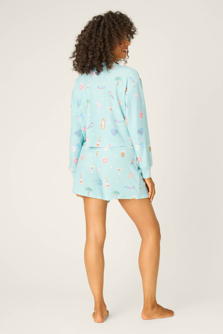 A person with curly hair stands barefoot, facing away, wearing the P.J. Salvage Boyfriend Boxer Ornament Print Shorts Set. The pajama set is light blue with colorful drink and palm tree patterns, against a plain, light-colored background.