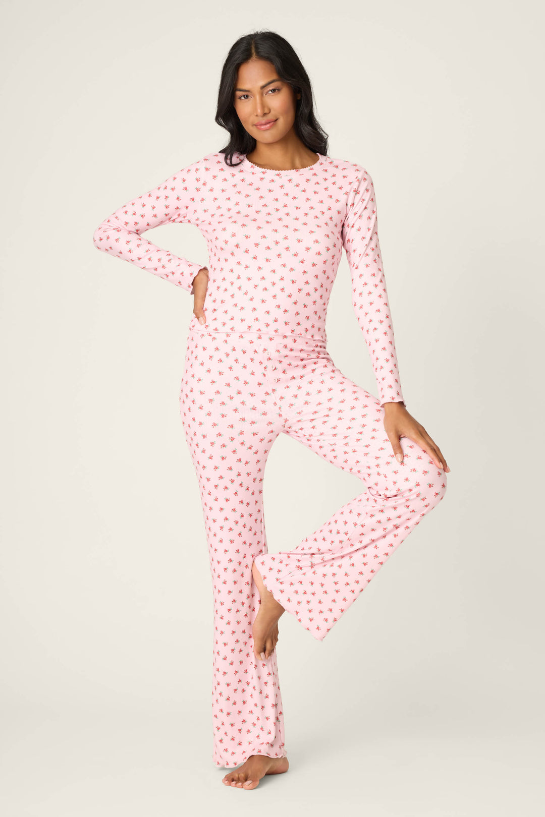 A person stands on one leg in the P.J. Salvage Crazy For The Holidays Ditsy Floral Print Pants Set, featuring a matching long-sleeve top and flared pants with a pink floral pattern, against a plain white background.