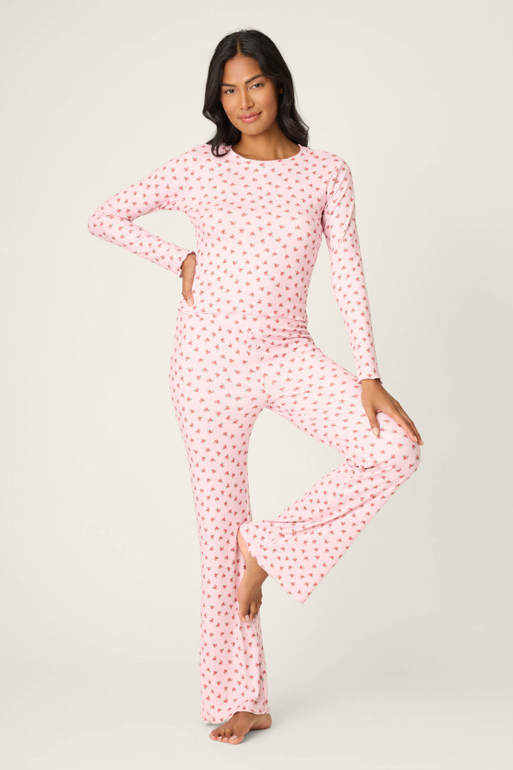 A person stands on one leg in the P.J. Salvage Crazy For The Holidays Ditsy Floral Print Pants Set, featuring a matching long-sleeve top and flared pants with a pink floral pattern, against a plain white background.