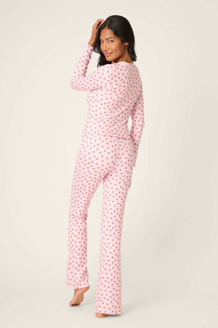 A woman stands barefoot, facing away and looking over her shoulder in P.J. Salvage's Crazy For The Holidays Ditsy Floral Print Pants Set, featuring a pink floral pattern, against a plain light background.