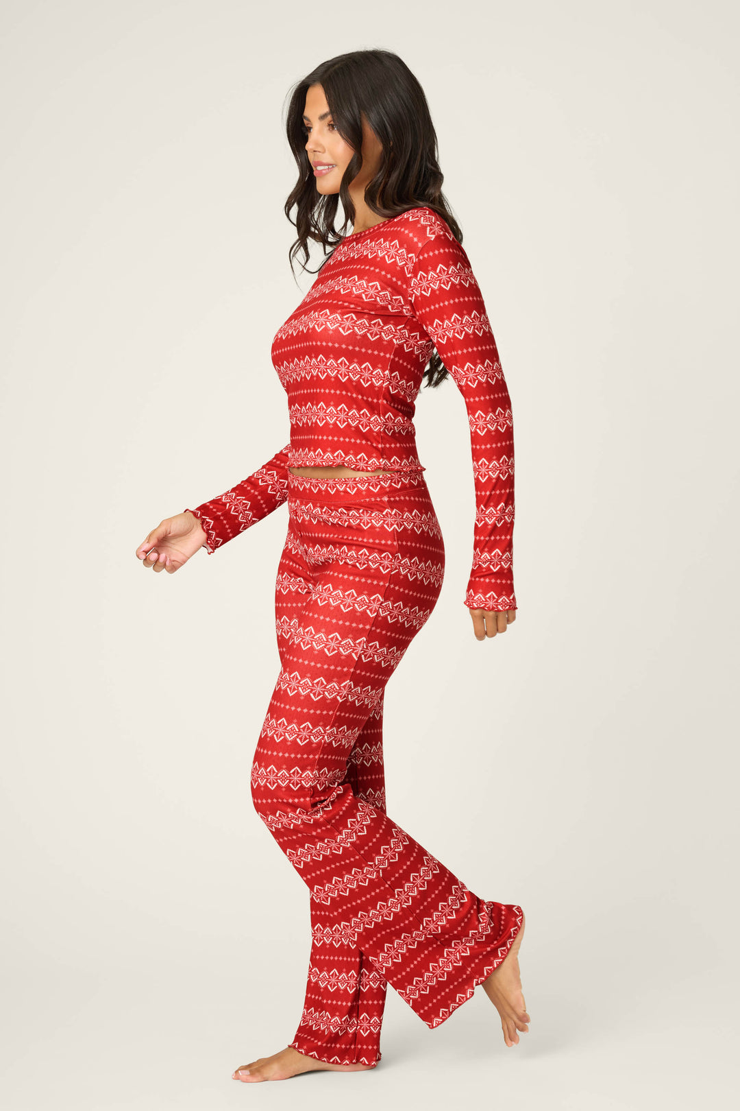 A woman with long dark wavy hair walks barefoot in the P.J. Salvage Crazy For The Holidays Fairisle Print Pants Set, featuring a red pointelle long-sleeve top and full-length pants with a white Fair Isle motif, against a plain light background.