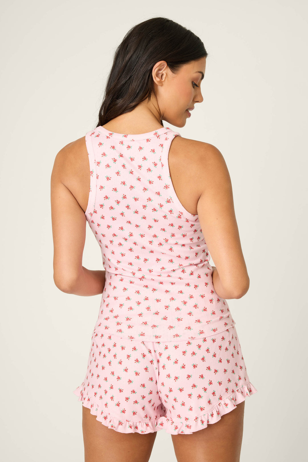 A woman with long dark hair faces away from the camera wearing the P.J. Salvage Crazy For The Holidays Ditsy Floral Print Tank Top, featuring a light pink hue with a red pattern and ruffled hems, against a plain light background.