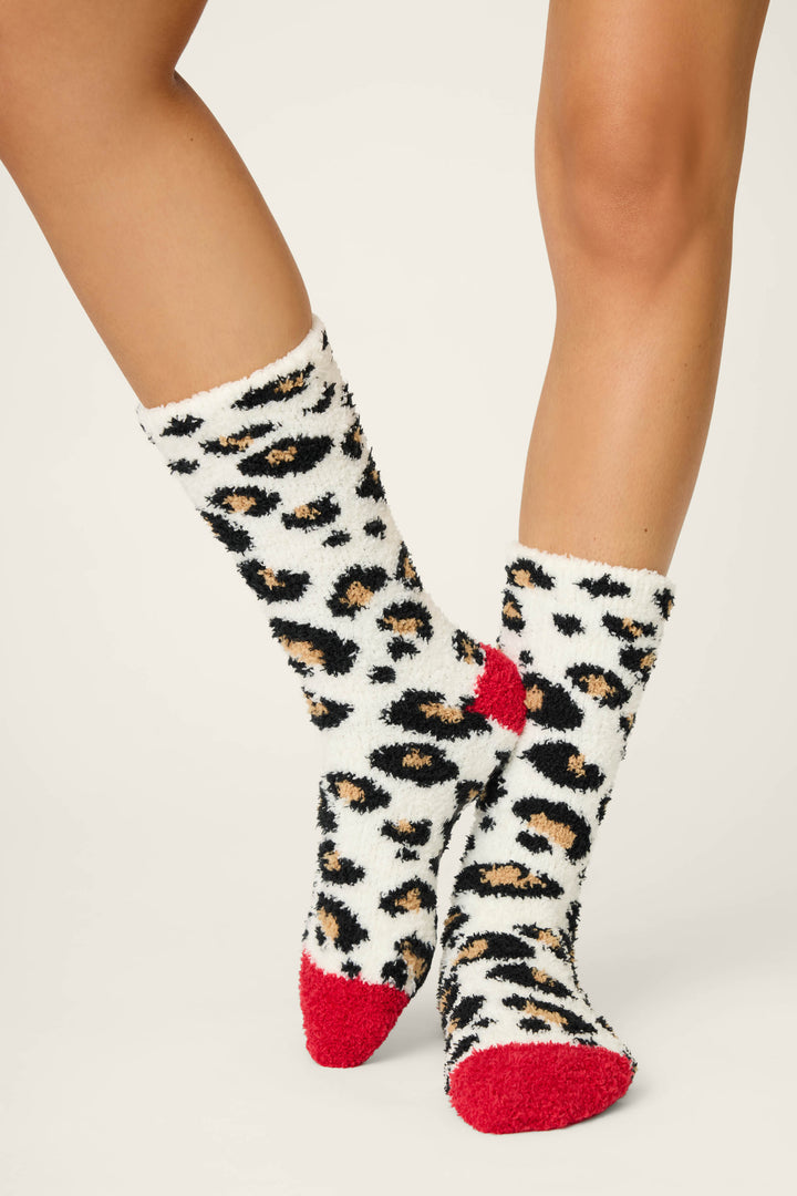 A person stands on a light background, feet crossed at the ankles, wearing P.J. Salvage LEOPARD SOCKS HOLIDAY DREAMS—fuzzy white crew socks with black and tan leopard spots plus bright red heels and toes.