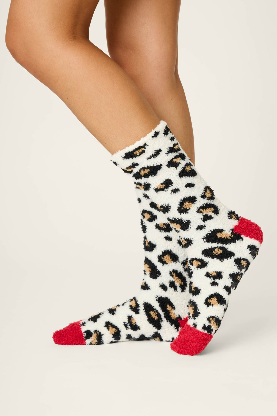 A person wears P.J. Salvage LEOPARD SOCKS HOLIDAY DREAMS—fuzzy white socks with black and tan leopard spots, plus solid red toes and heels—standing on a plain light background.