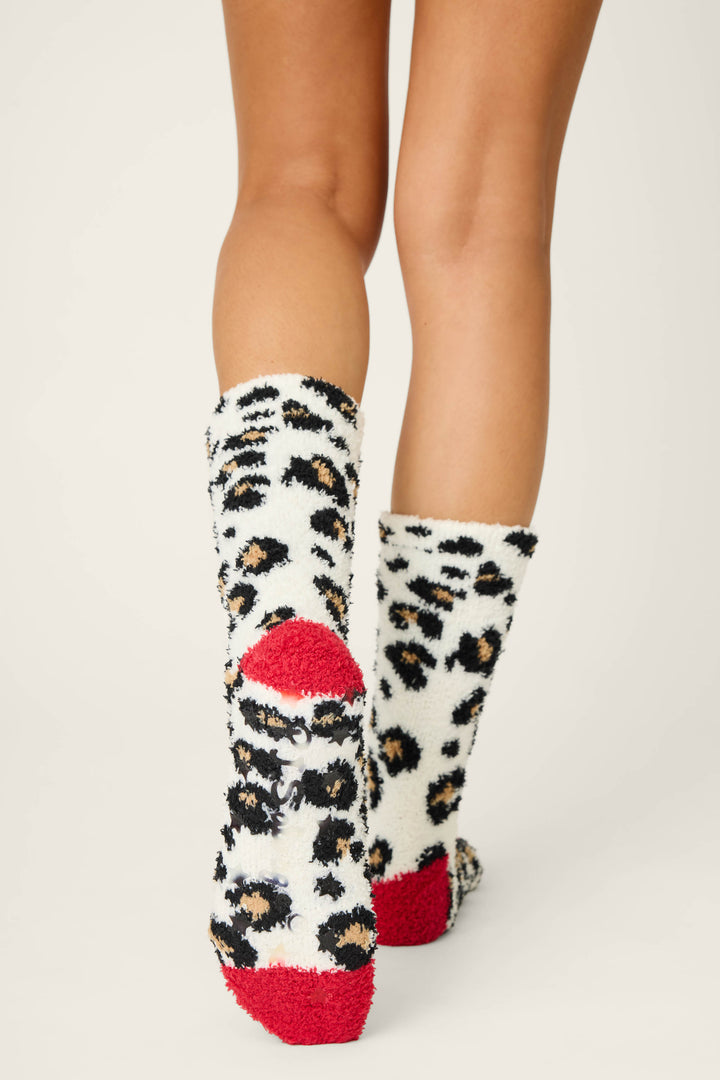 A person stands on a light surface wearing P.J. Salvage LEOPARD SOCKS HOLIDAY DREAMS: fuzzy white socks with black and tan leopard print spots, plus red heels and toes. Only their lower legs and feet are visible.