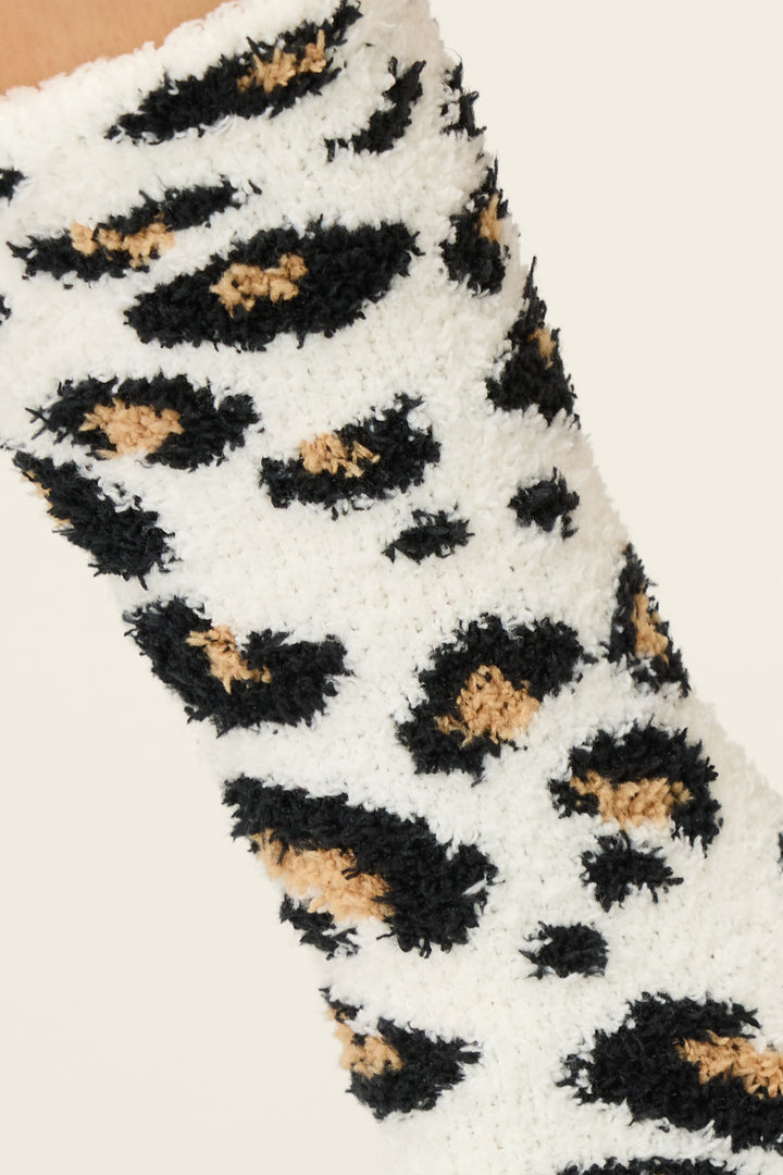 A close-up of the P.J. Salvage Holiday Dreams Leopard Print Crew Socks with Grippers, featuring a fuzzy white fabric and a black and tan leopard print, displayed on a plain light background.