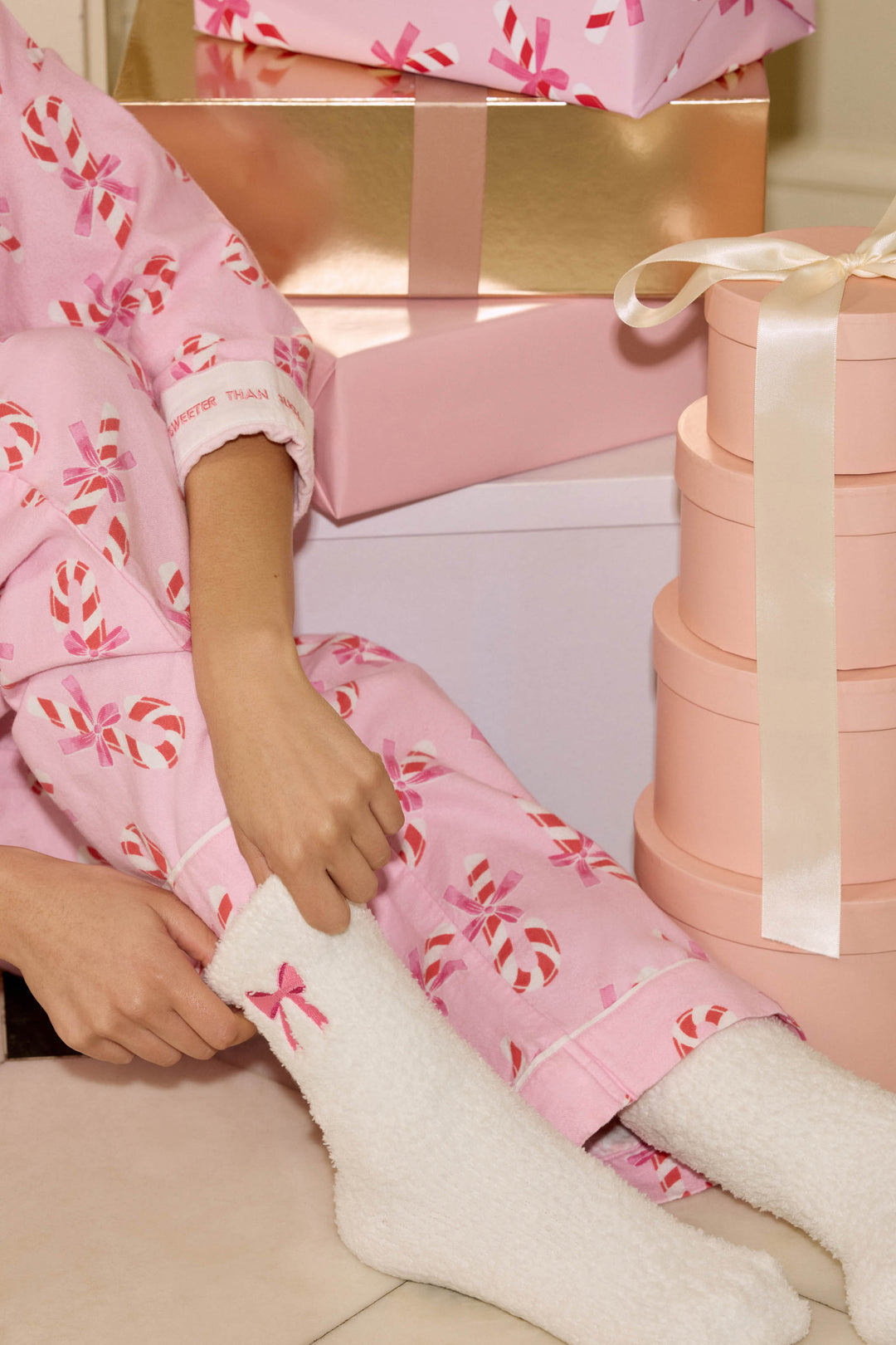 A person in pink pajamas adjusts their P.J. Salvage Bow Print Crew Socks with Grippers, featuring a pink bow. Pink and gold gift boxes are stacked in the background.