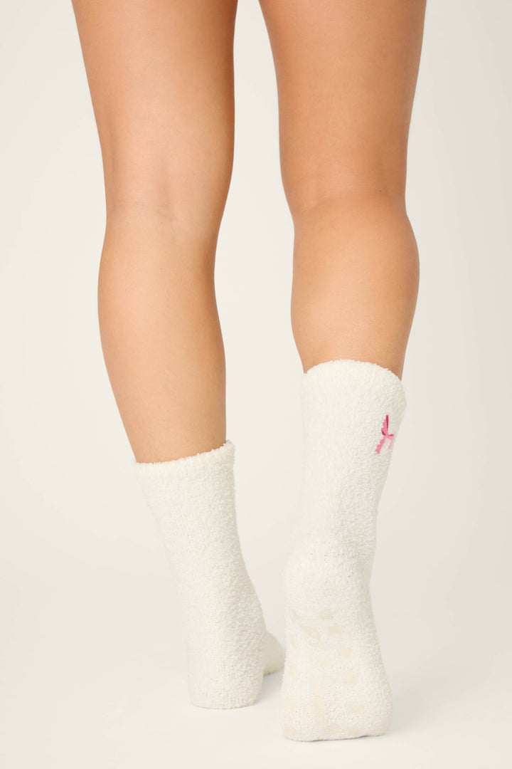 A person stands on tiptoe from behind, wearing P.J. Salvage Bow Print Crew Socks with Grippers featuring a small pink ribbon on the back. The white fuzzy socks are shown against a plain, light background.