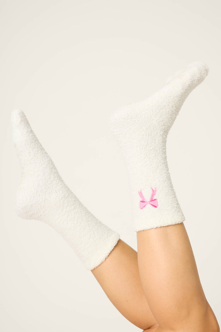 A pair of feet wearing P.J. Salvage Bow Print Crew Socks with Grippers, featuring a fuzzy white fabric and a small pink bow near the ankle, raised in the air against a plain light background.