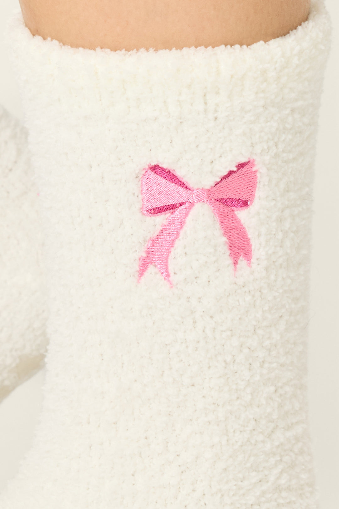A close-up of P.J. Salvage Bow Print Crew Socks with Grippers, showcasing a fuzzy white design and an embroidered pink bow near the top.