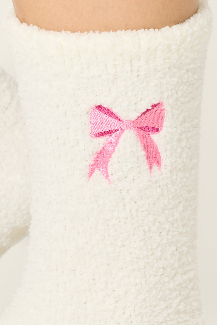 A close-up of P.J. Salvage Bow Print Crew Socks with Grippers, showcasing a fuzzy white design and an embroidered pink bow near the top.