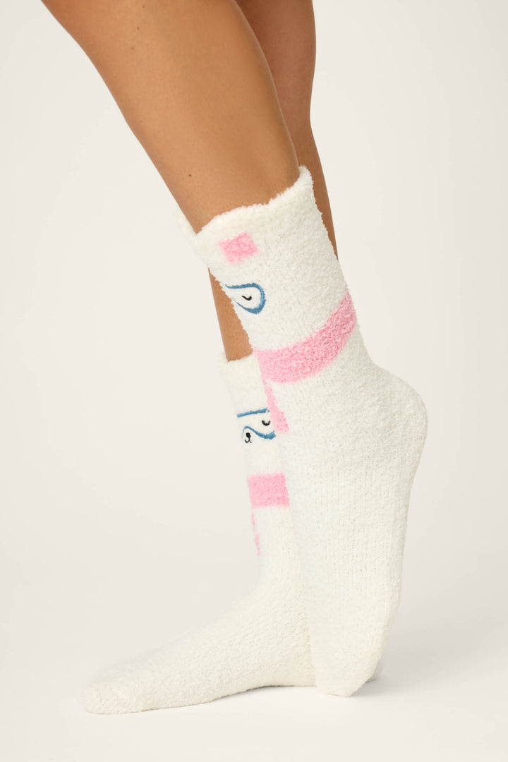 A person stands on their toes against a plain light background, wearing P.J. Salvage BEAR SOCKS FAM JAM BEARLY AWAKE white fuzzy socks with pink stripes and blue cartoon eye designs.