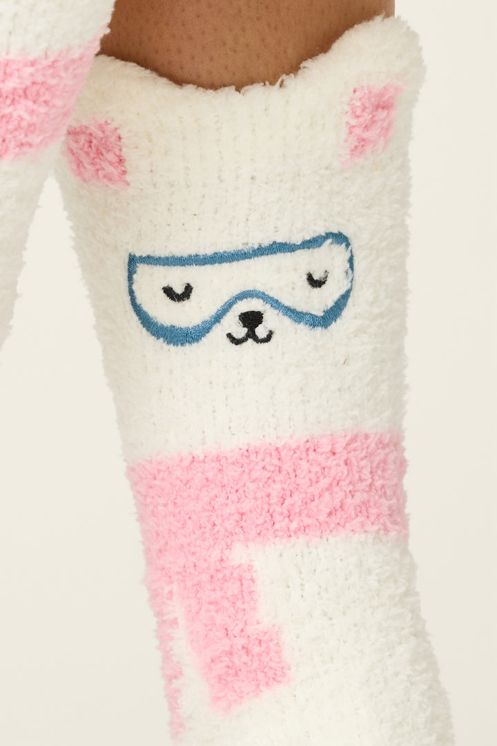 A close-up of P.J. Salvage’s Chill Out Bunny Print Crew Socks with Grippers, worn on a leg. The fuzzy white sock features pink patches and a cute bunny face design with blue goggles and pink ears.