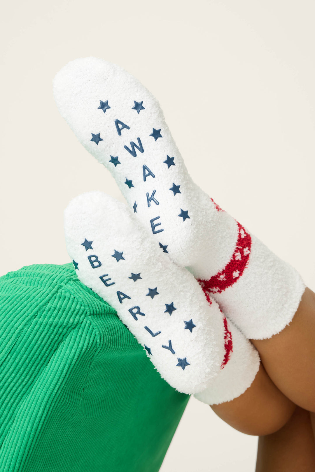 A person wearing P.J. Salvage BEAR SOCKS FAM JAM BEARLY AWAKE, featuring white fuzzy fabric with blue stars and “AWAKE/BARELY” on the soles, rests their feet on a green surface while dressed in red and white pants.