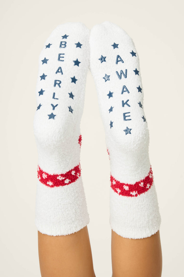 Someone wears P.J. Salvage BEAR SOCKS FAM JAM BEARLY AWAKE: white fuzzy socks with red and white star bands, and blue "BEARLY AWAKE" text with stars on the soles.