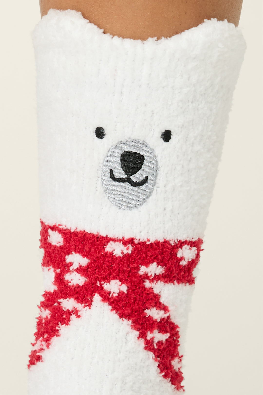 A close-up of P.J. Salvage Fam Jam Bear Print Crew Socks with Grippers, showing a fuzzy white sock with a bear face near the top and a red scarf with white polka dots below the face.