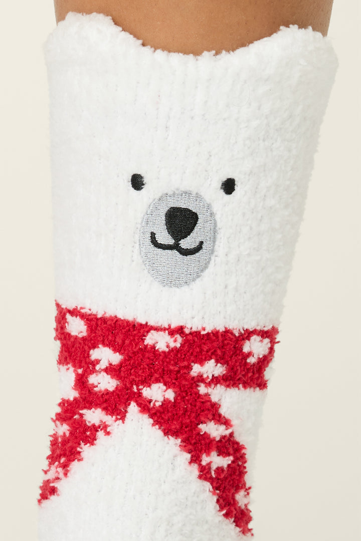 A close-up of P.J. Salvage Fam Jam Bear Print Crew Socks with Grippers, showing a fuzzy white sock with a bear face near the top and a red scarf with white polka dots below the face.