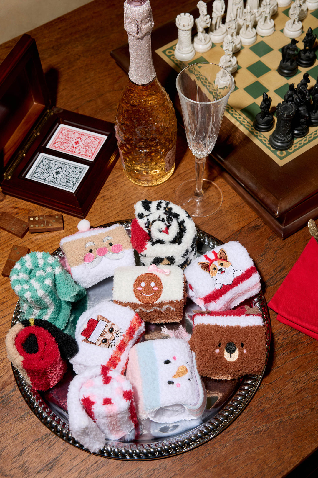 A wooden table displays P.J. Salvage CAT SOCKS FELINE FESTIVE in a silver tray, surrounded by a bottle of sparkling drink, a glass, red napkins, two decks of cards, dice, and a chessboard.