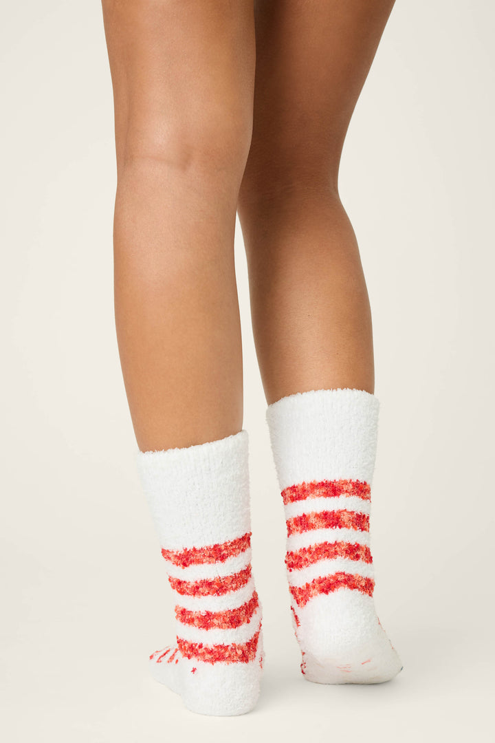 A person faces away from the camera, displaying the back of their legs and feet clad in P.J. Salvage CAT SOCKS FELINE FESTIVE—white fuzzy socks with red stripes—against a plain light background.