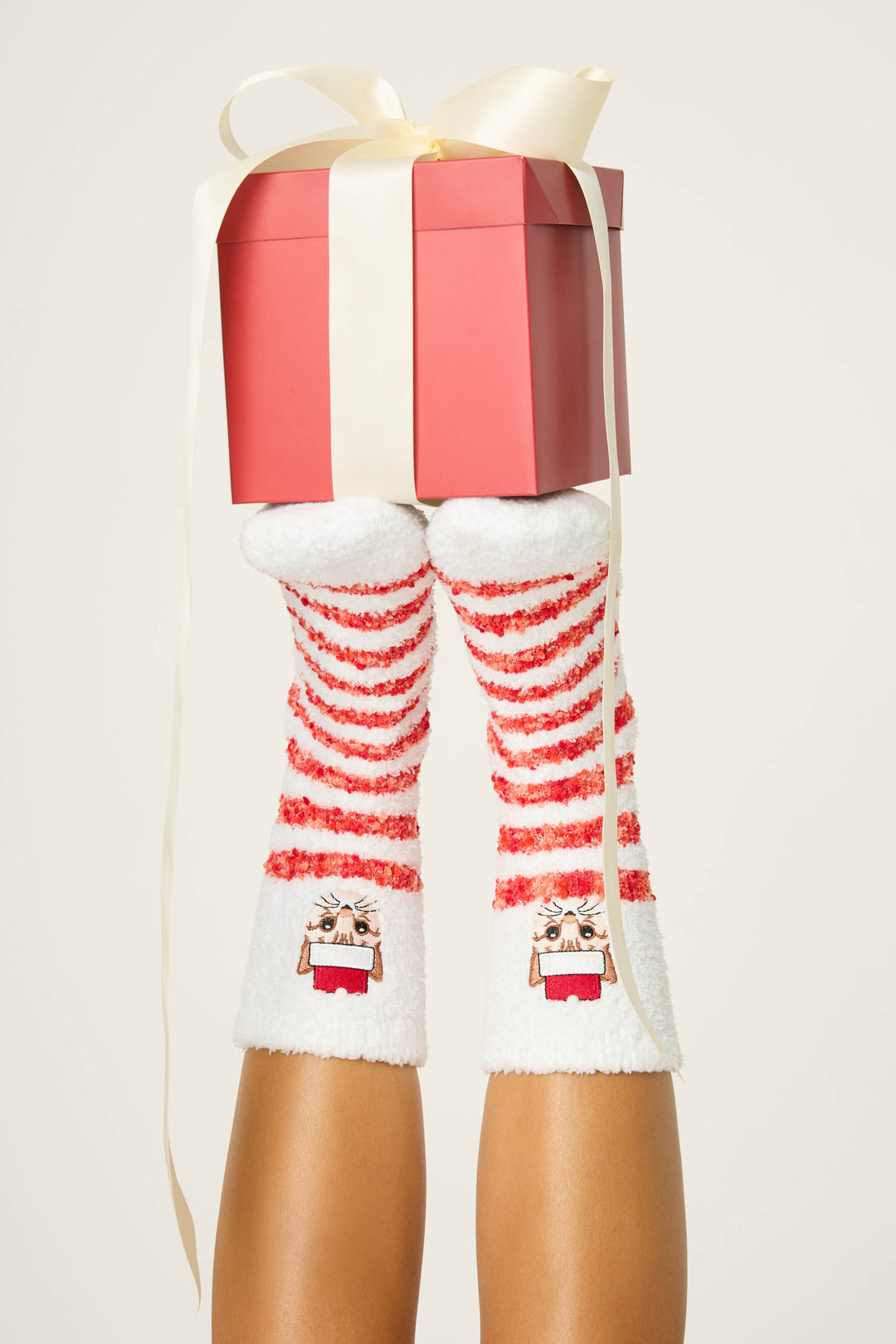 A person’s legs are raised vertically, wearing P.J. Salvage CAT SOCKS FELINE FESTIVE—red and white striped fuzzy socks with a cat design. The feet balance a pink gift box tied with a cream ribbon against a light background.