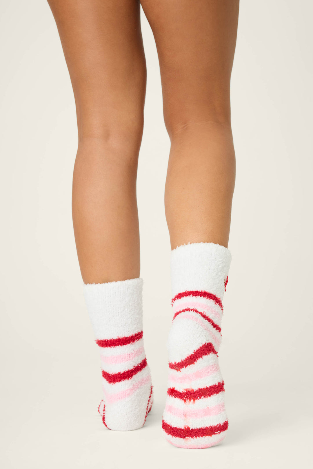 A person stands with legs visible from mid-thigh down, wearing P.J. Salvage's DOG SOCKS PEACE & PAWLIDAYS—fuzzy white socks featuring red and pink stripes. The background is plain and light-colored.