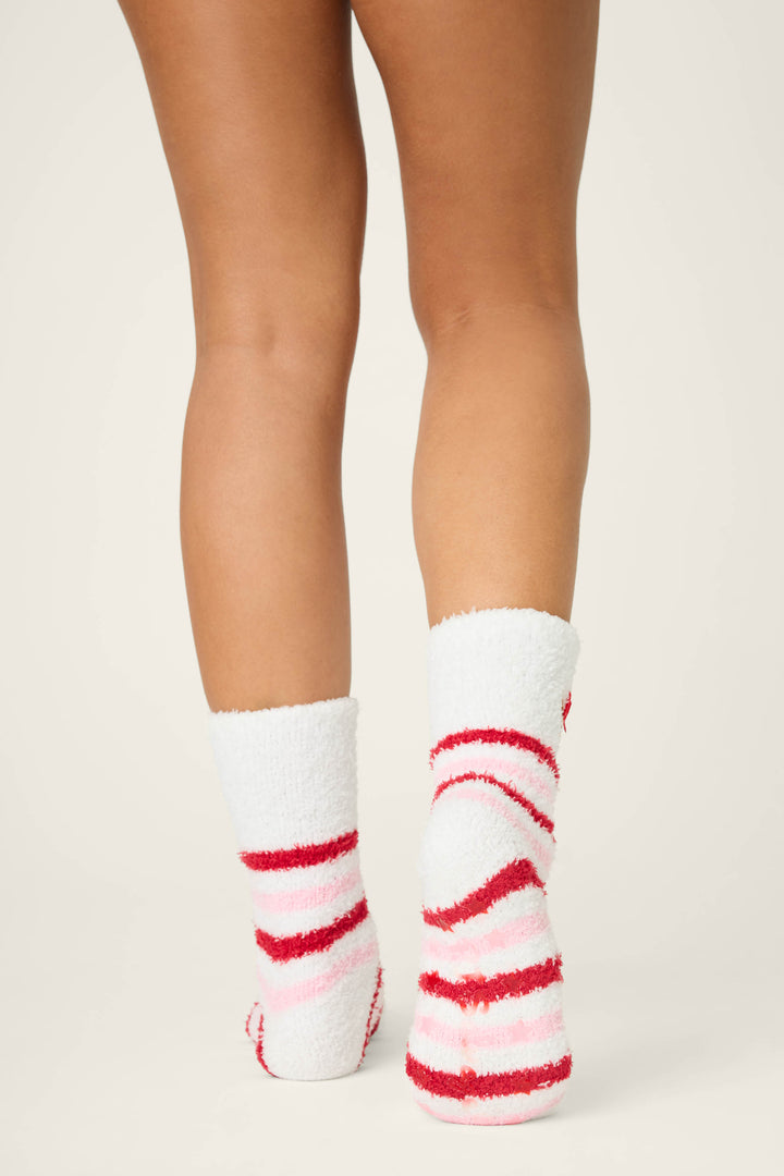 A person stands with legs visible from mid-thigh down, wearing P.J. Salvage's DOG SOCKS PEACE & PAWLIDAYS—fuzzy white socks featuring red and pink stripes. The background is plain and light-colored.