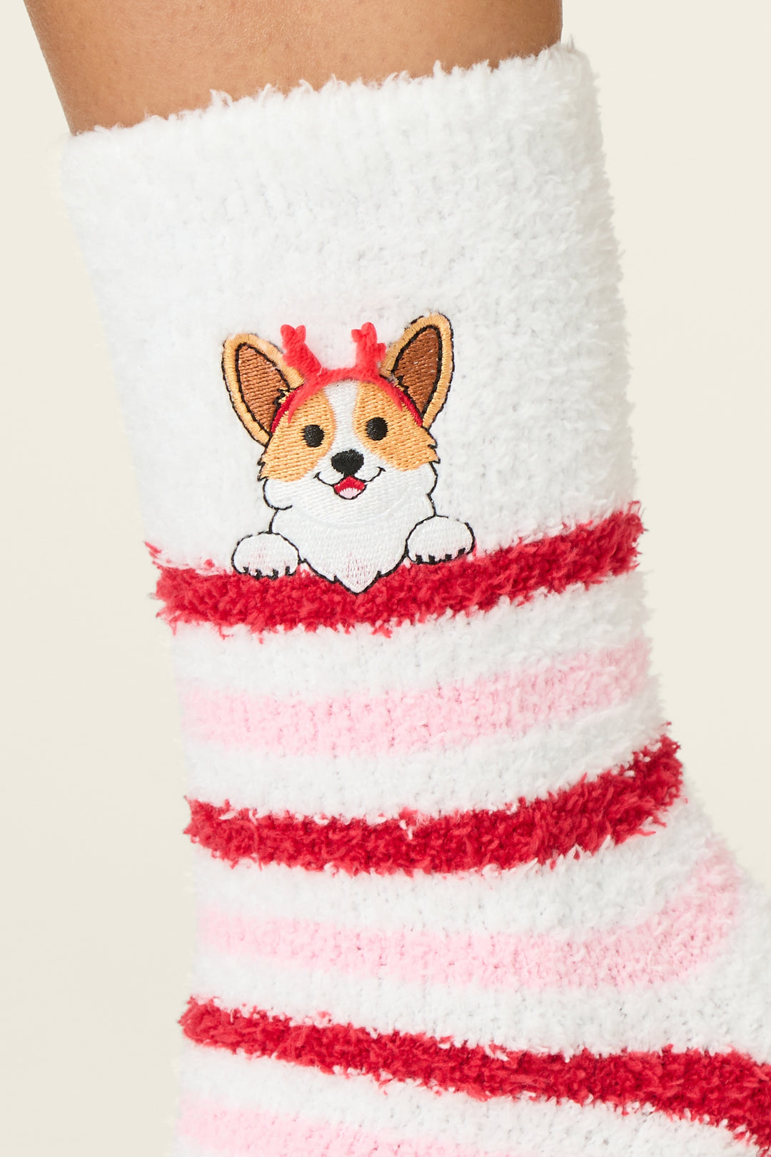A close-up of P.J. Salvage Peace & Pawlidays Corgi Print Crew Socks with Grippers, featuring fuzzy white fabric, red and pink stripes, and an embroidered corgi dog with red reindeer antlers.