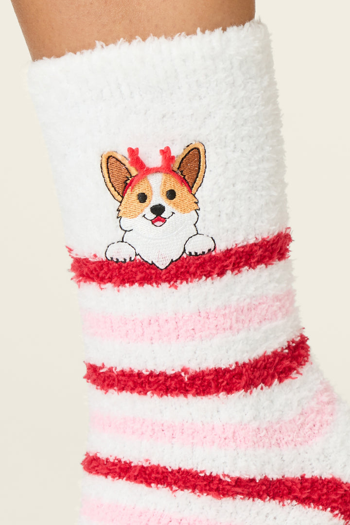 A close-up of P.J. Salvage Peace & Pawlidays Corgi Print Crew Socks with Grippers, featuring fuzzy white fabric, red and pink stripes, and an embroidered corgi dog with red reindeer antlers.