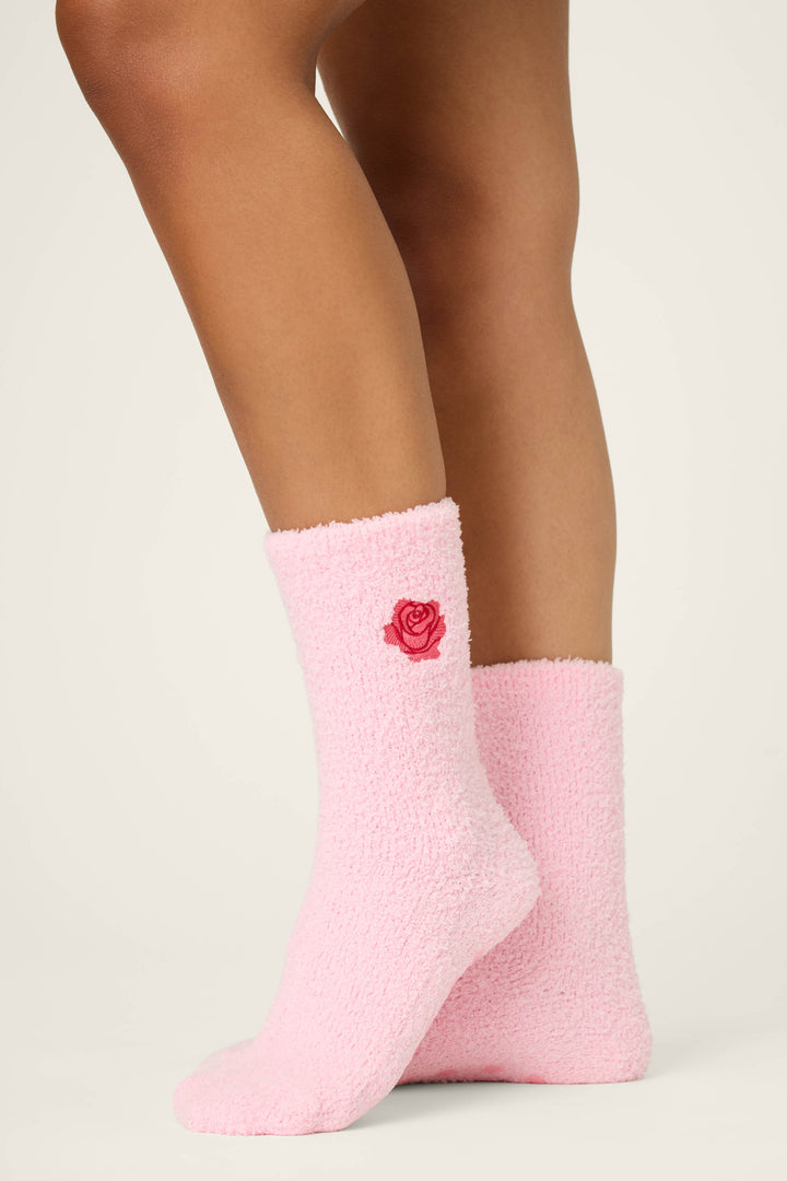 A person models P.J. Salvage’s ROSE SOCKS CRAZY FOR THE HOLIDAYS—pink fuzzy socks with a small red rose near the top—standing with one foot slightly ahead against a light background.