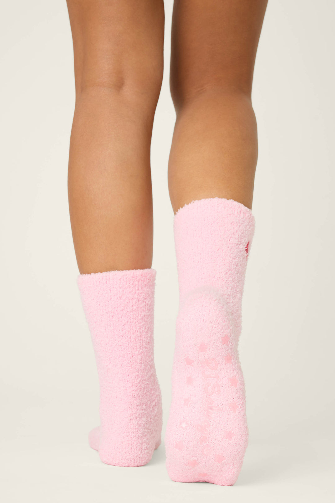 A person, shown from behind, stands on a light surface wearing P.J. Salvage's ROSE SOCKS CRAZY FOR THE HOLIDAYS—knee-length pink fuzzy socks with non-slip grips. Only their lower legs and feet are visible.