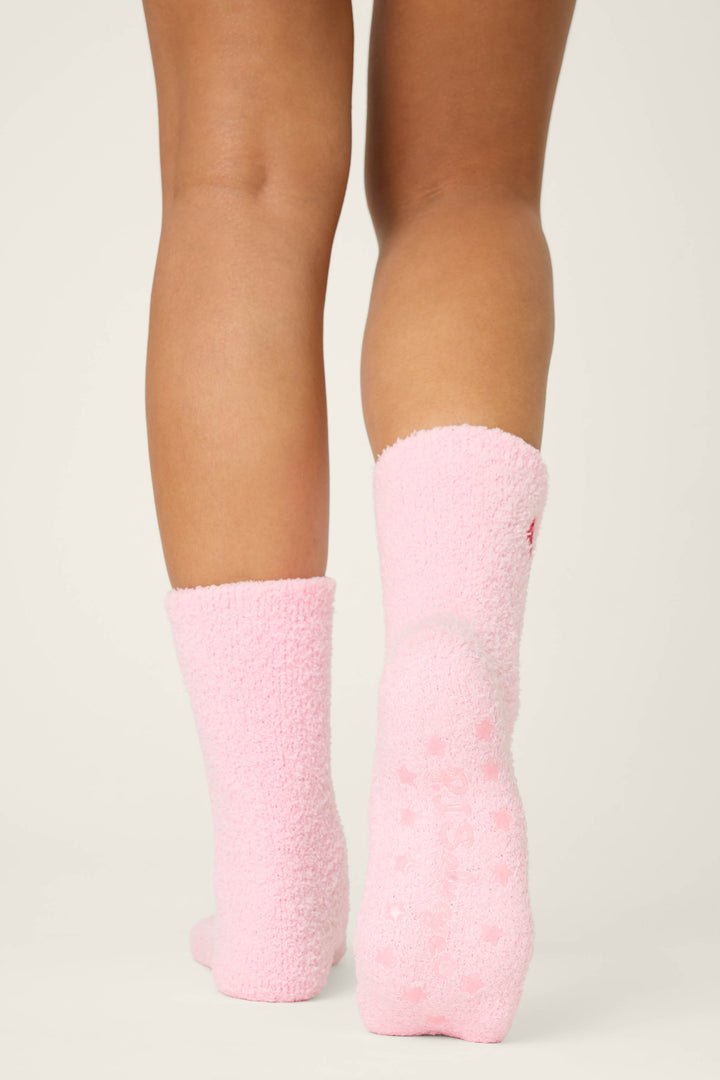 A person, shown from behind, stands on a light surface wearing P.J. Salvage's ROSE SOCKS CRAZY FOR THE HOLIDAYS—knee-length pink fuzzy socks with non-slip grips. Only their lower legs and feet are visible.