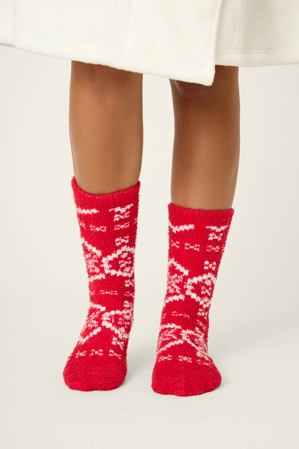 A person in a white robe stands on a light surface, wearing P.J. Salvage FAIRISLE SOCKS CRAZY FOR THE HOLIDAYS—red fuzzy socks featuring a white winter-themed pattern.