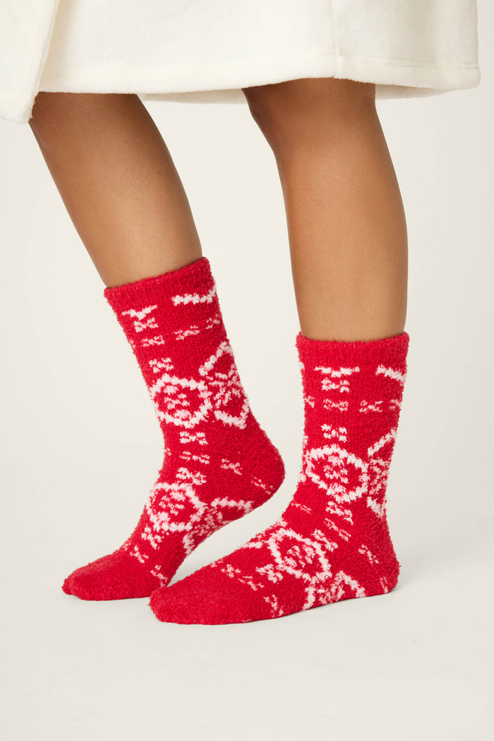 A person in a white garment stands on a light background, wearing P.J. Salvage FAIRISLE SOCKS CRAZY FOR THE HOLIDAYS—red fuzzy socks with white winter-themed patterns.