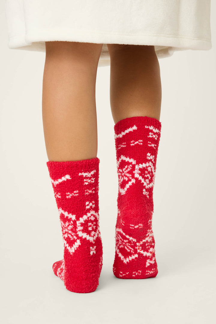 A person in a white garment stands with their back visible, wearing P.J. Salvage FAIRISLE SOCKS CRAZY FOR THE HOLIDAYS—red socks with white snowflake patterns—against a plain, light background.