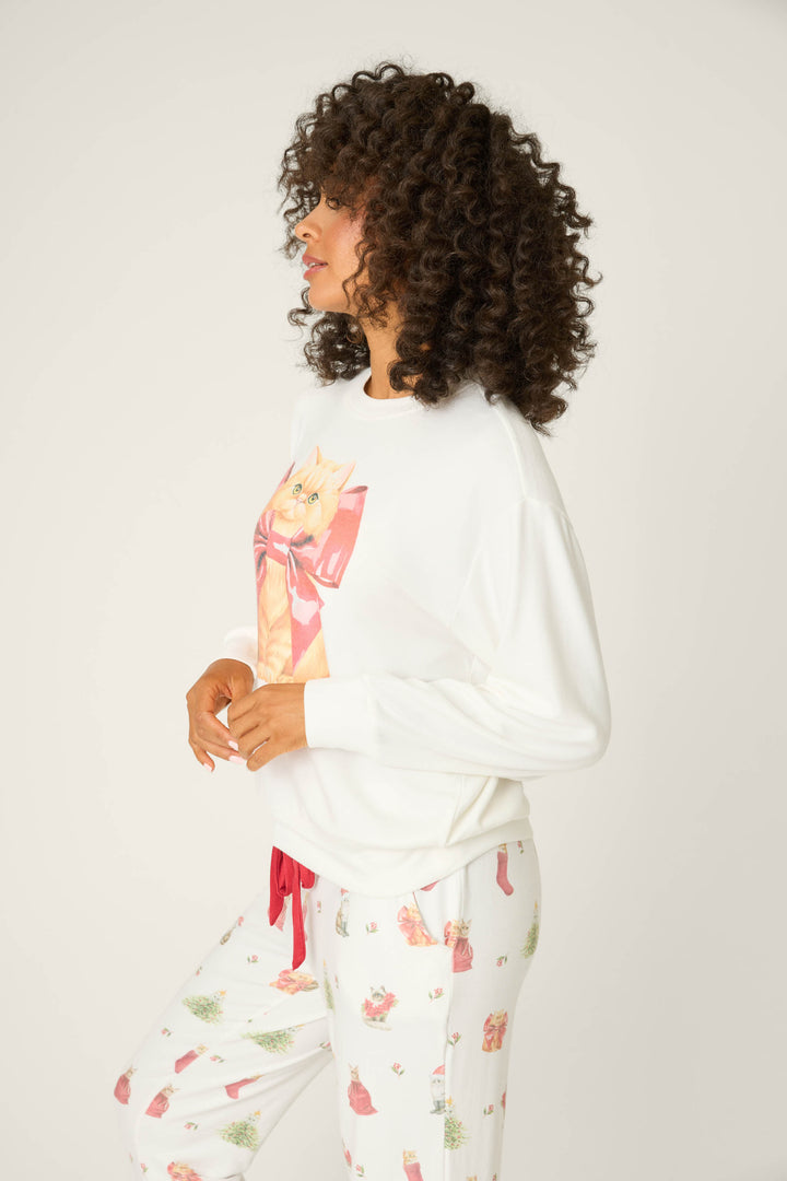A person with curly hair stands sideways, wearing the P.J. Salvage Feline Festive Cat Print Sweater and white holiday-themed pajama pants, posed against a plain background.
