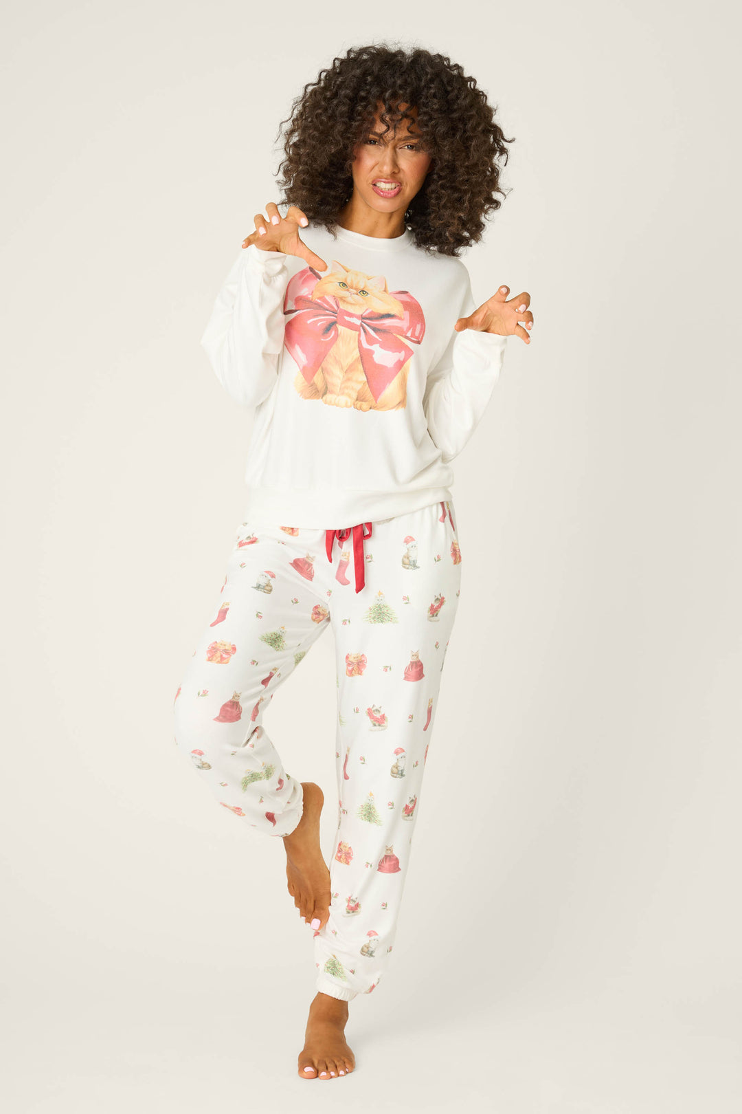 A curly-haired woman stands barefoot on one leg, playfully raising her hands like claws. She wears the P.J. Salvage Feline Festive Cat Print Sweater and matching pajama pants decorated with cats, teapots, and cupcakes.