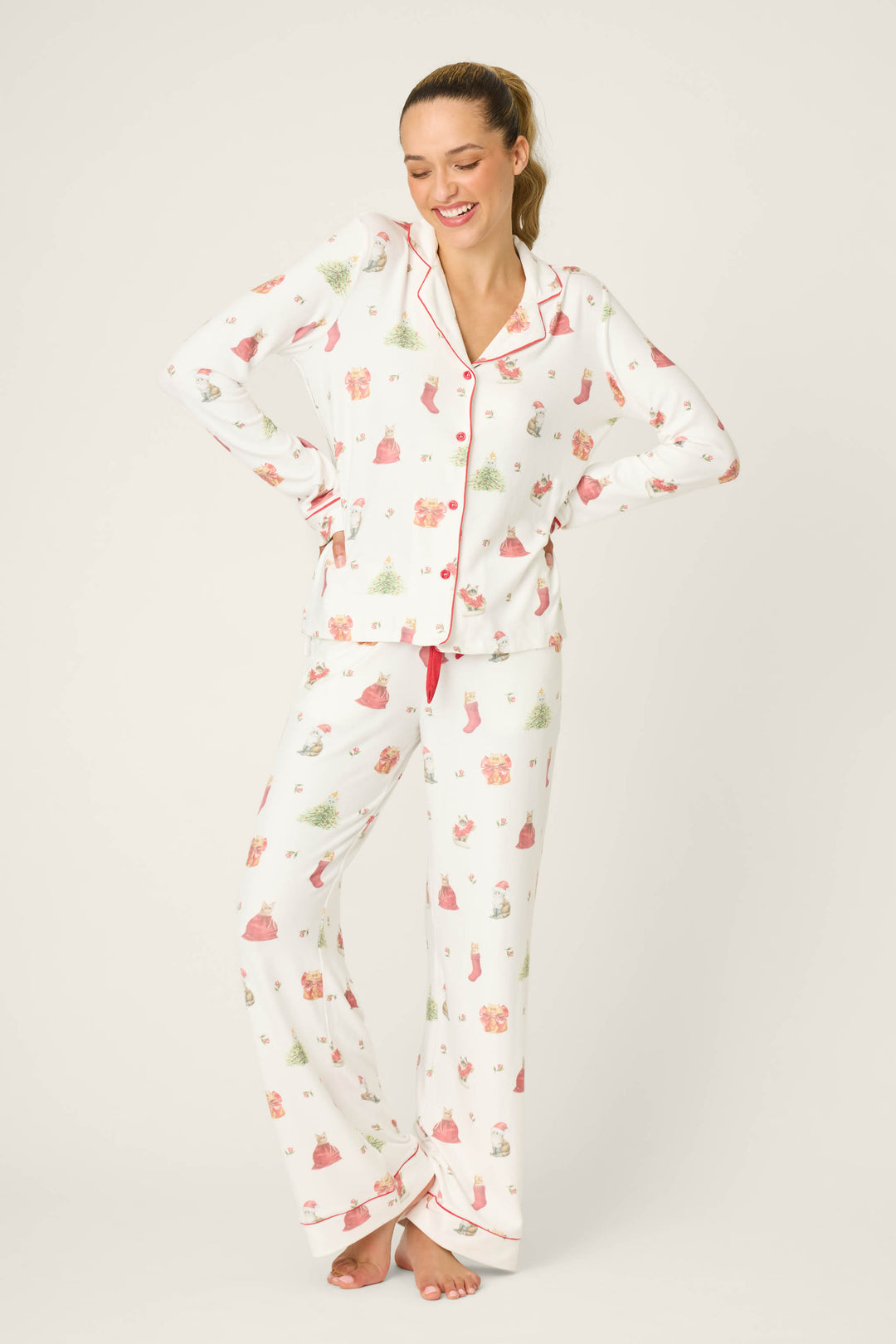A woman stands barefoot, smiling with hands on hips, wearing the P.J. Salvage Feline Festive Cat Print PJ Set—white pajama pants and a long-sleeve top with red piping and holiday cat prints—against a plain background.