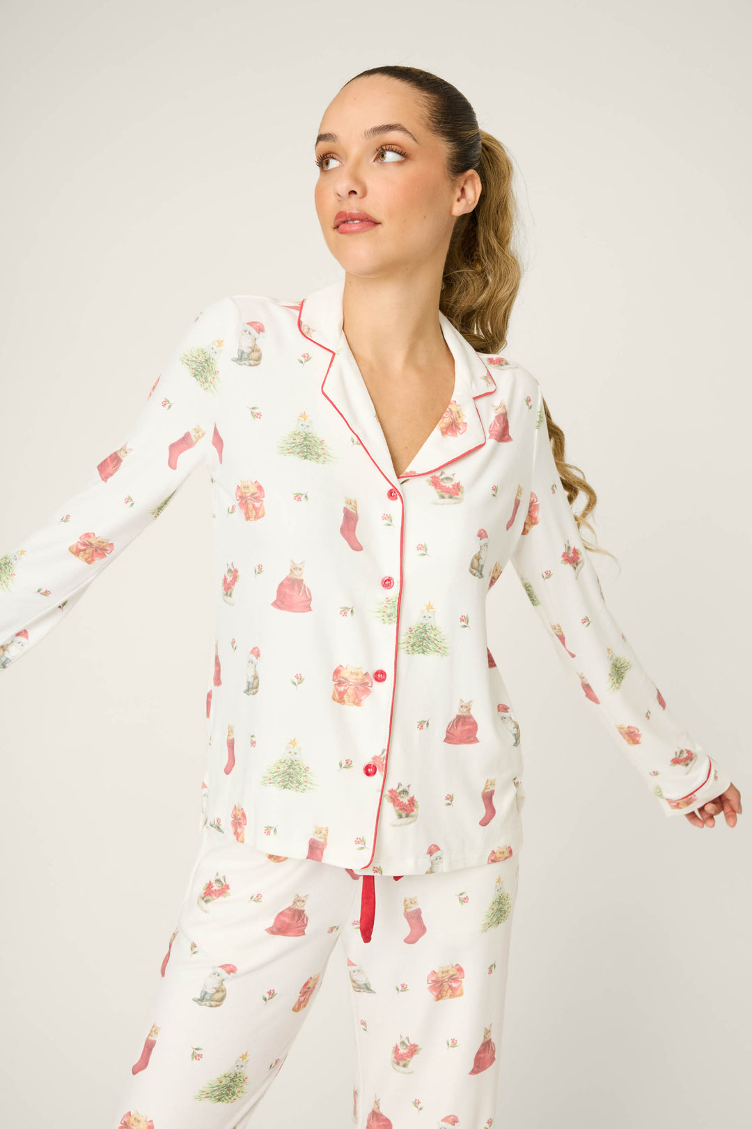A woman with long, light brown hair in a ponytail wears the P.J. Salvage Feline Festive Cat Print PJ Set, featuring white pajamas with red piping and cat holiday prints, standing against a plain light background and looking to the side.