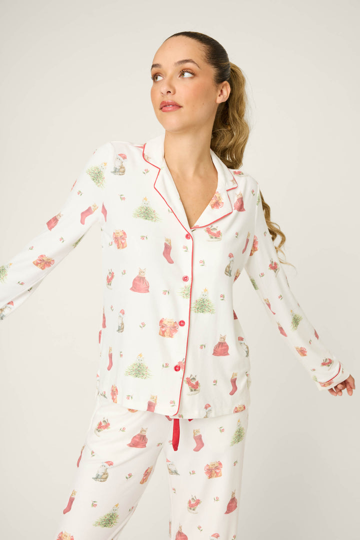 A woman with long, light brown hair in a ponytail wears the P.J. Salvage Feline Festive Cat Print PJ Set, featuring white pajamas with red piping and cat holiday prints, standing against a plain light background and looking to the side.