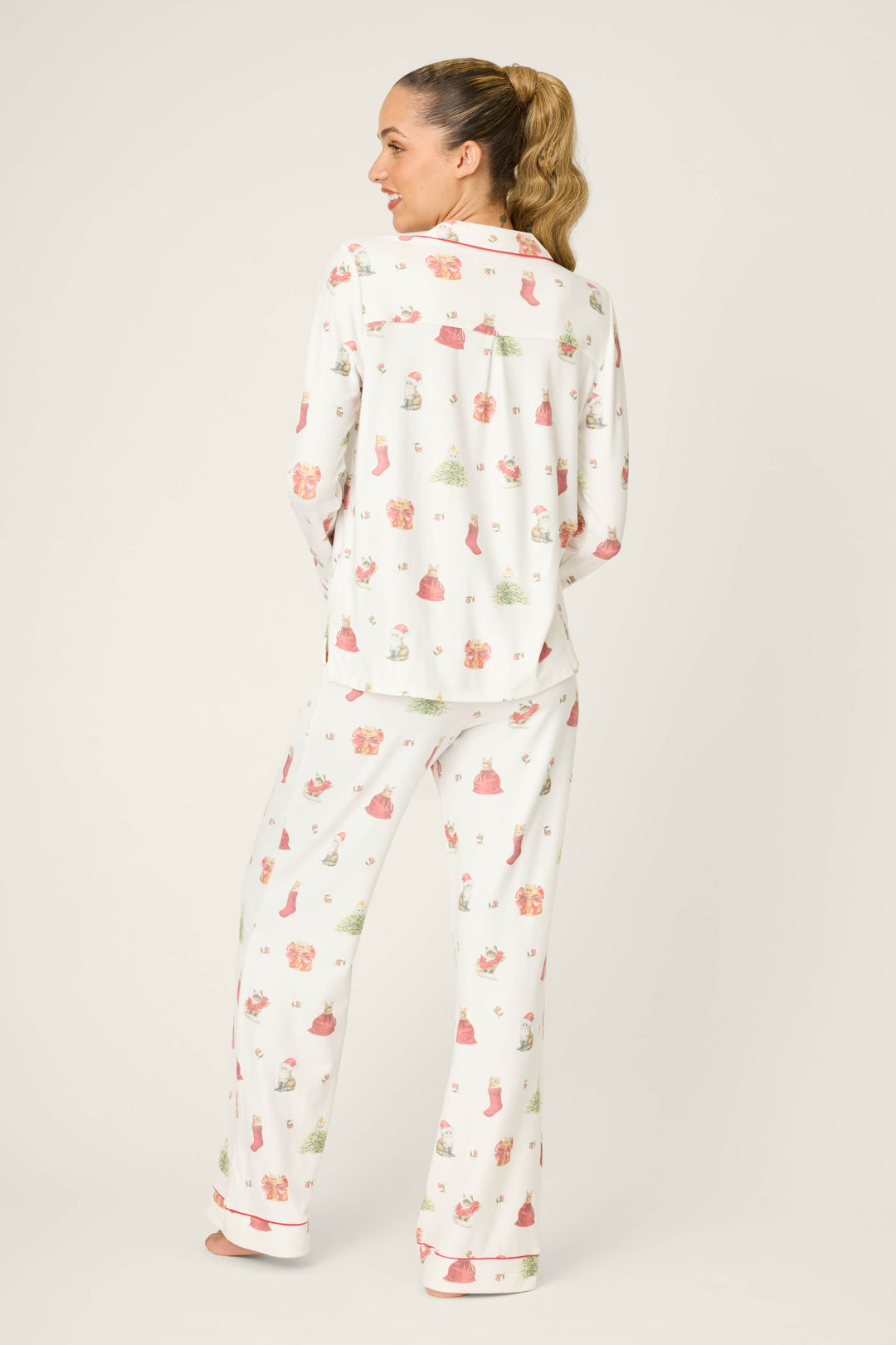 A woman with her hair in a ponytail models the P.J. Salvage Feline Festive Cat Print PJ Set, featuring a relaxed-fit, long-sleeved top and pants with white fabric and festive pink and green cat prints, shown on a plain background.