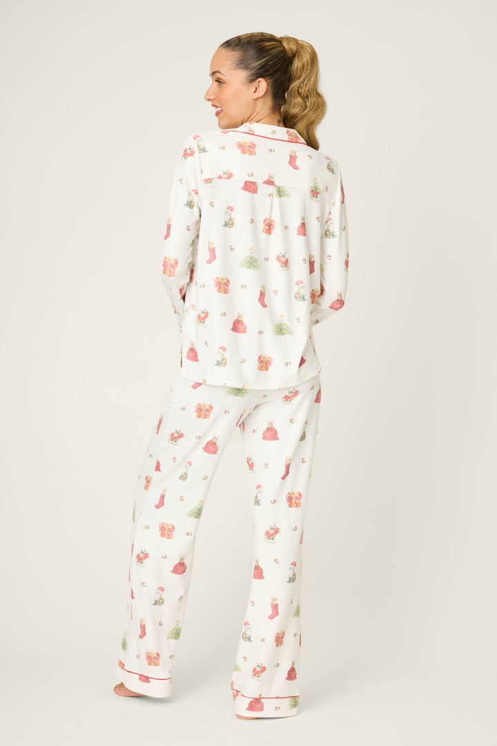 A woman with her hair in a ponytail models the P.J. Salvage Feline Festive Cat Print PJ Set, featuring a relaxed-fit, long-sleeved top and pants with white fabric and festive pink and green cat prints, shown on a plain background.