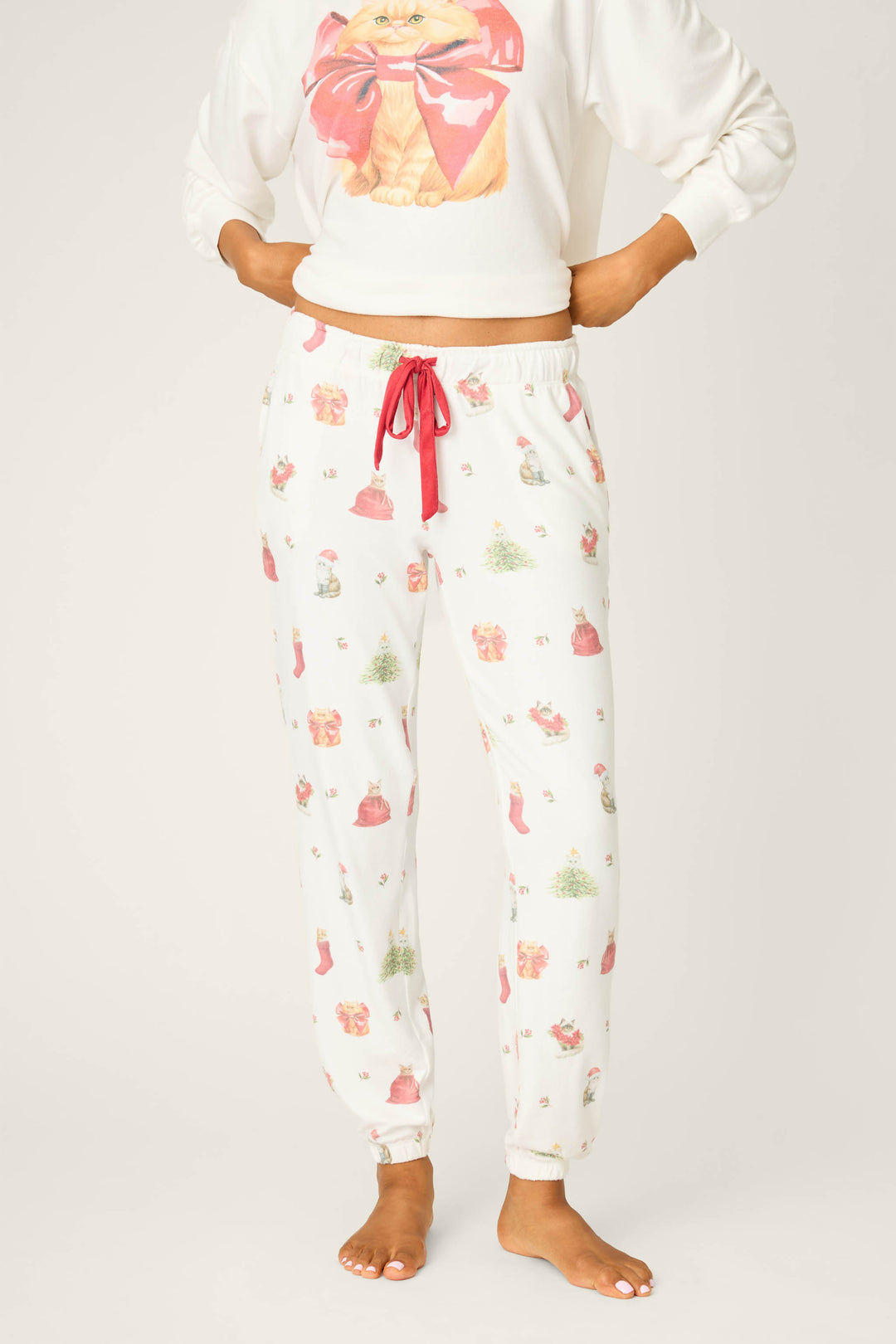 A person wears P.J. Salvage's Feline Festive Cat Print Banded Pants, featuring gifts, Christmas trees, and stockings, paired with a sweatshirt with a cat holding a red bow, standing barefoot against a plain background.